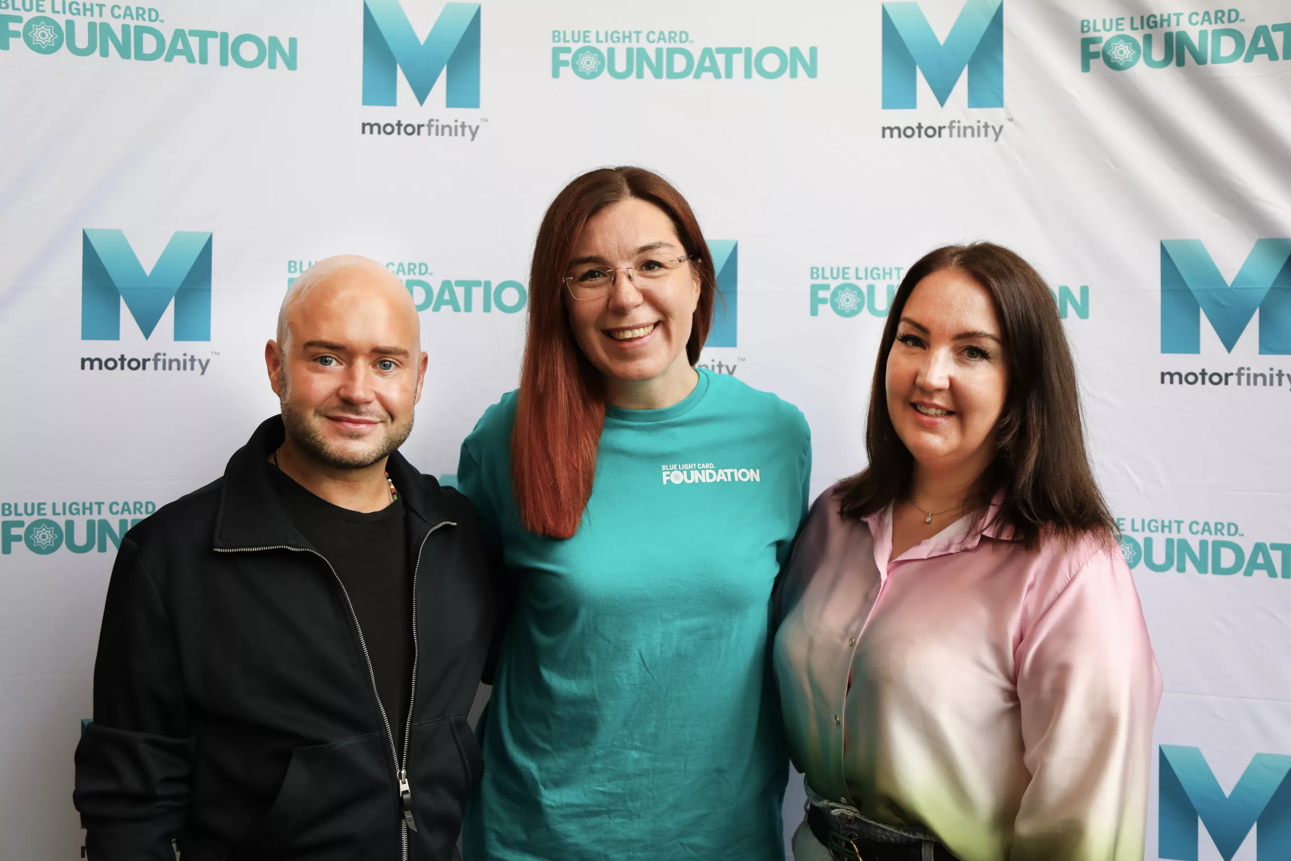 A picture of two Motorfinity staff and Blue Light Card Foundation's fundraising manager. They are standing in front of a backdrop showing both logos.